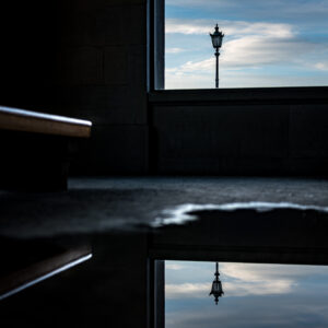 Strassenlaterne als Silhouette gegen blauen Himmel, eingerahmt von dunkler Architektur und gespiegelt in einer Pfütze – Featured Image FotoTipp Bildgestaltung in der Fotografie photomundo.