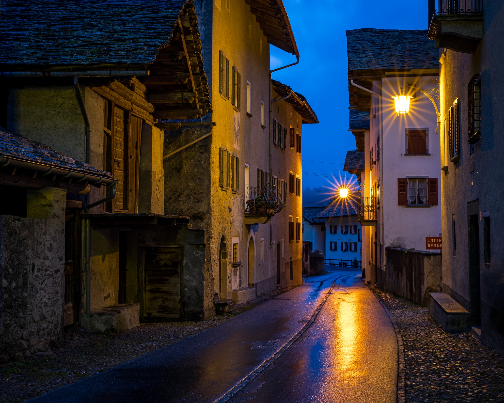 Blaue Stunde fotografieren – nasse Dorfgasse mit Strassenlampenlicht und Spiegelung auf dem Asphalt