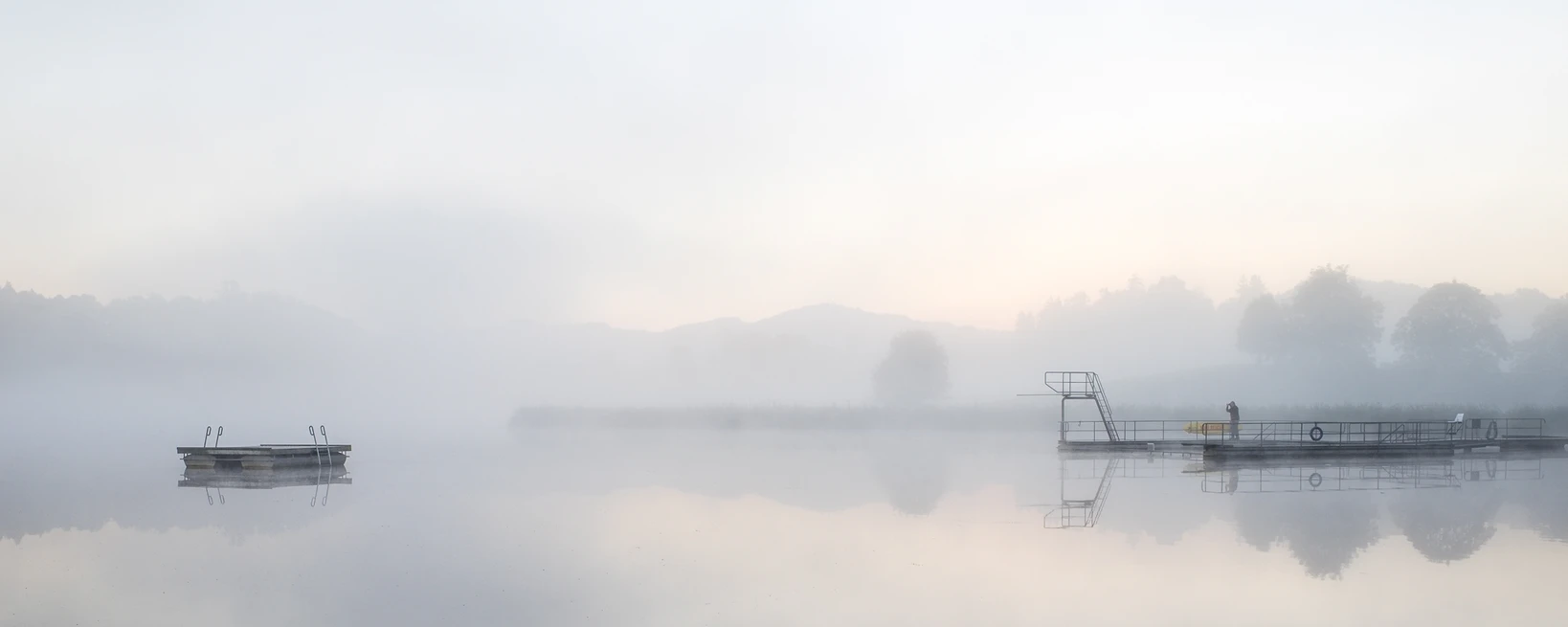 Morgennebel über einem stillen See mit zwei Schwimmstegen und ihrer Spiegelung im Wasser, eine stille Gestalt im Gegenlicht, ruhige Bilder entstehen in solchen Momenten