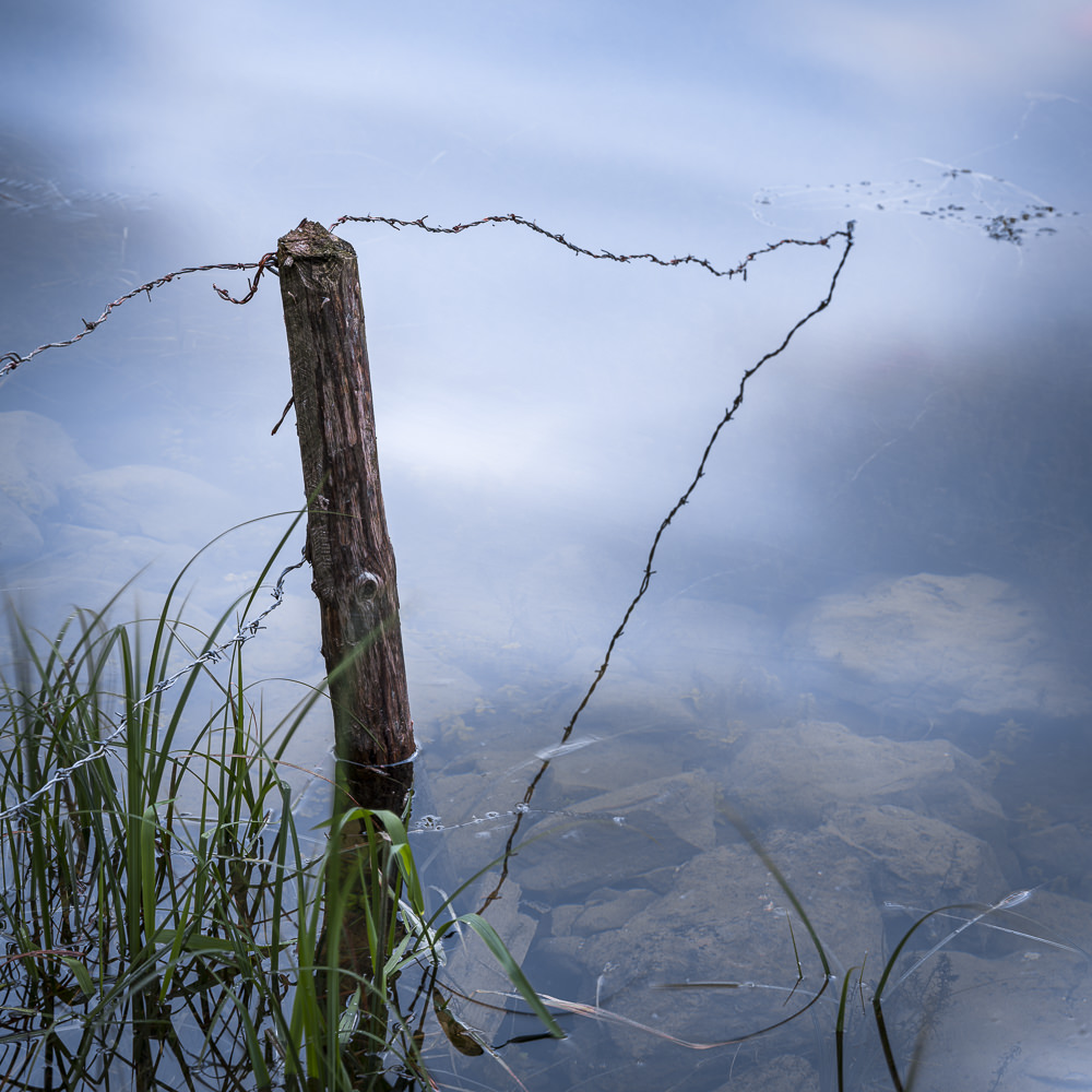 Alter Holzpfahl mit geschwungenem Draht am Ufer eines klaren Sees.